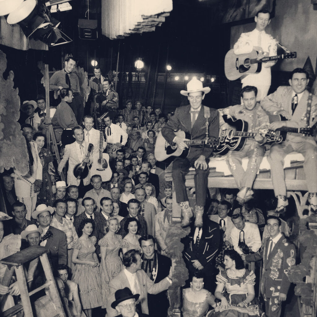 Large group of people in western wear, cowboy hats, boots, holding guitars.